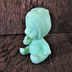 May include: A small, green, sitting baby figurine made of soap. The baby is wearing a hooded hat and has a sweet expression on its face.