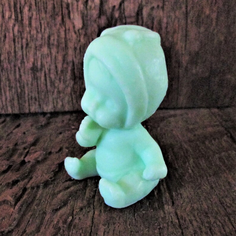 May include: A small, green, sitting baby figurine made of soap. The baby is wearing a hooded hat and has a sweet expression on its face.