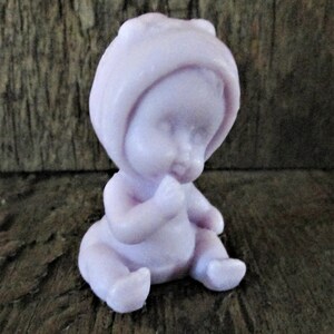 May include: A small, lavender-colored baby doll figurine with a bonnet, sitting with its hands clasped in front of it.