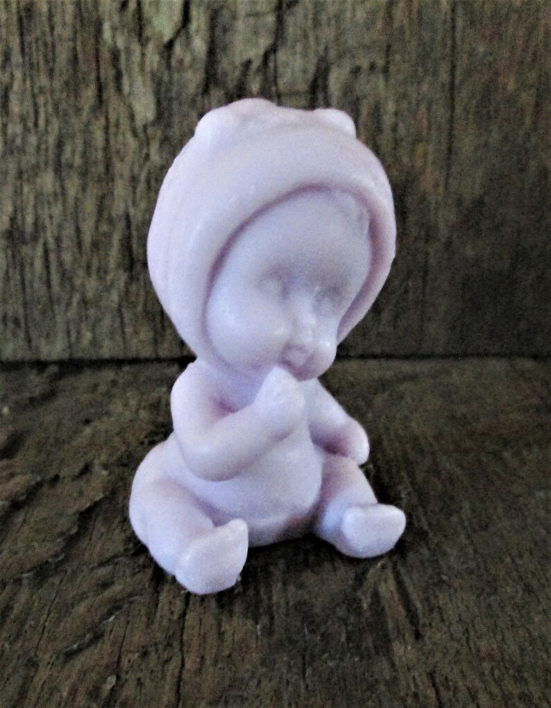 May include: A small, lavender-colored baby doll figurine with a bonnet, sitting with its hands clasped in front of it.