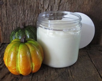 May include: A clear jar containing a creamy white substance, likely a lotion or cream, is positioned beside two decorative pumpkins. One pumpkin is orange and yellow, the other green. The background is a rustic wooden surface.