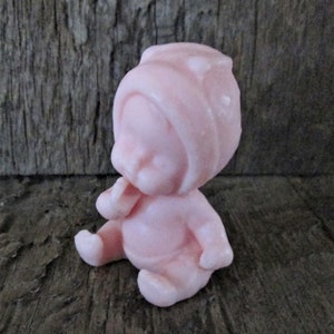 May include: A small pink baby doll figurine wearing a pink hat. The doll is sitting with its hands clasped in front of it.