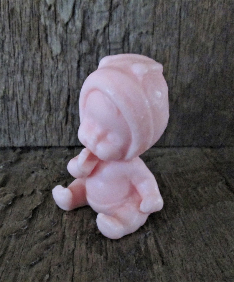 May include: A small pink baby doll figurine wearing a pink hat. The doll is sitting with its hands clasped in front of it.