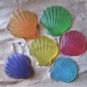 May include: Six colorful seashell-shaped soaps, each with a glittery finish. The soaps are arranged on a sandy surface. The colors of the soaps are orange, green, pink, yellow, blue, and purple.