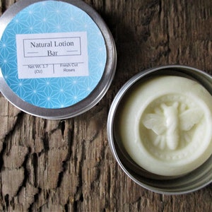 Peut inclure: Une petite boîte en argent avec une étiquette géométrique bleu et blanc qui indique "Natural Lotion Bar - Net Wt 1.7 (Oz) Fresh Cut Roses". La boîte contient une barre de lotion blanche avec un motif d'abeille.