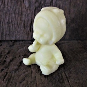 May include: A small, pale yellow plastic figurine of a baby wearing a hooded hat. The baby is sitting with its hands clasped in front of its face.