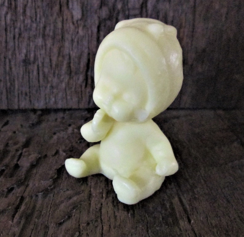 May include: A small, pale yellow plastic figurine of a baby wearing a hooded hat. The baby is sitting with its hands clasped in front of its face.