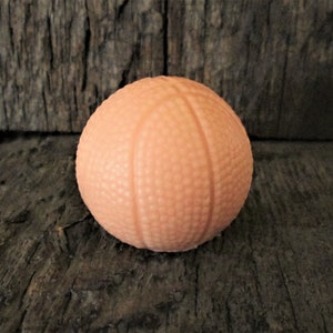May include: A small, peach-colored ball with a textured surface and vertical lines, resembling a miniature basketball. The ball is set against a dark, textured wooden background.