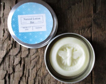 May include: A small tin container with a white lid and a blue and white label that reads "Natural Lotion Bar" and "Net Wt. 1.7 oz Gardenia". The container is open and a white lotion bar with a bee design is visible inside.