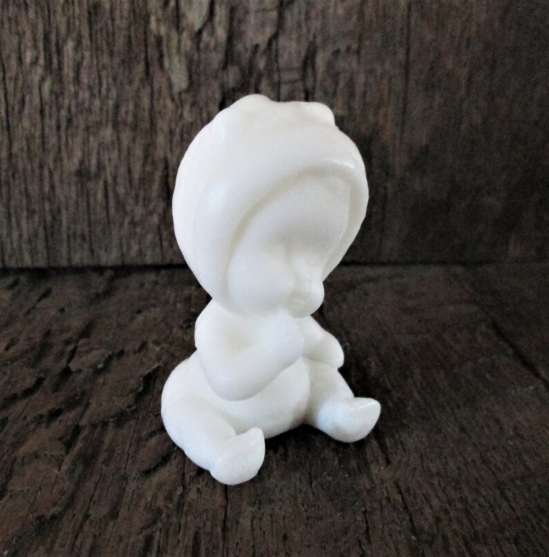 May include: A white ceramic figurine of a baby wearing a hooded onesie. The baby is sitting with its hands clasped in front of its face.