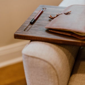 May include: A brown leather-bound journal with a ribbon bookmark sits on a wooden tray with a black pen. The tray is resting on a light beige couch.