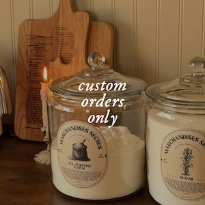 Puede incluir: Dos tarros de vidrio con tapas, etiquetados como "Harina para todo uso" y "Azúcar", llenos de polvo blanco. Los tarros tienen etiquetas de estilo vintage y están colocados frente a tablas de cortar de madera y una vela encendida. El texto en el frasco dice "solo pedidos personalizados".