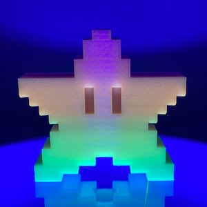 Op de afbeelding: Een 3D-geprint stervormig object met een pixelvormig ontwerp. De ster is gemaakt van een doorschijnend materiaal dat in het donker gloeit, met een kleurverloop van paars naar groen. De ster ligt op een blauw oppervlak.