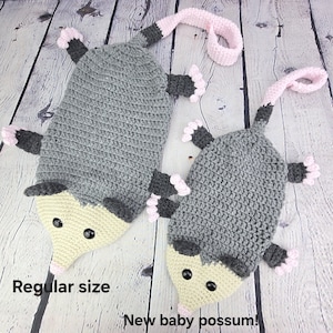 May include: Two crocheted possum plushies, one larger than the other. The larger possum is gray with a light yellow face and black eyes. The smaller possum is also gray with a light yellow face and black eyes. Both possums have pink ears and pink feet. The larger possum is labeled "Regular size" and the smaller possum is labeled "New baby possum!"