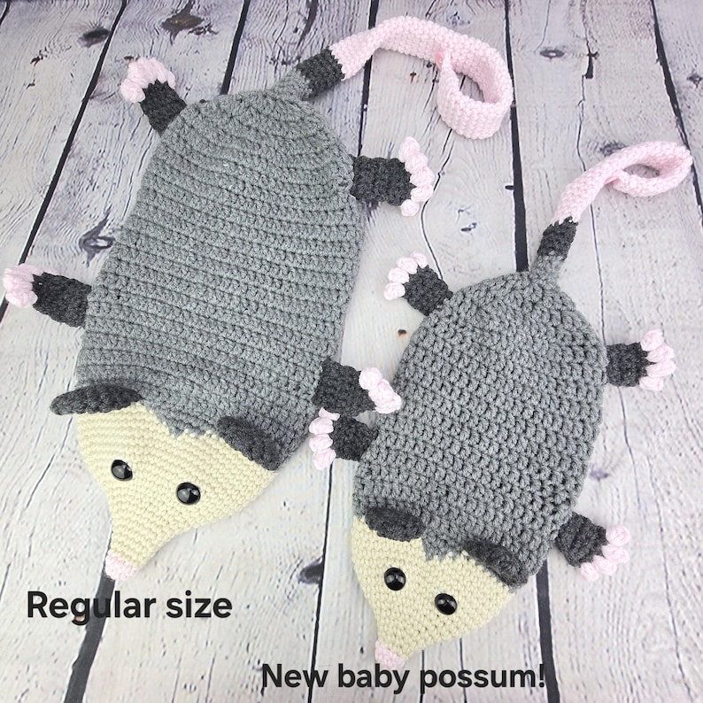 May include: Two crocheted possum plushies, one larger than the other. The larger possum is gray with a light yellow face and black eyes. The smaller possum is also gray with a light yellow face and black eyes. Both possums have pink ears and pink feet. The larger possum is labeled "Regular size" and the smaller possum is labeled "New baby possum!"