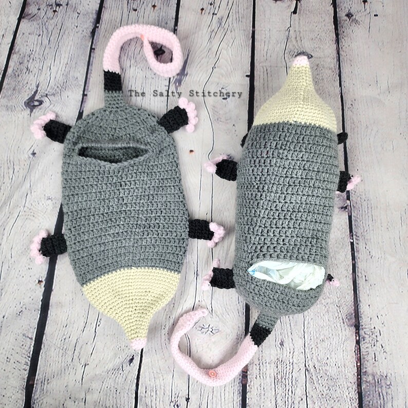 May include: Two crocheted gray and beige mole-shaped stuffed animals with pink accents. The animals are lying on a wooden surface. The text "The Salty Stitchery" is visible in the background.