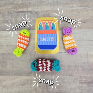 May include: A gold-colored rectangular tin labeled FIDGET FISH with three small, crocheted fish toys. One fish is orange and green, one is purple and white, and one is red, white, and blue. The word snap is printed around the items.