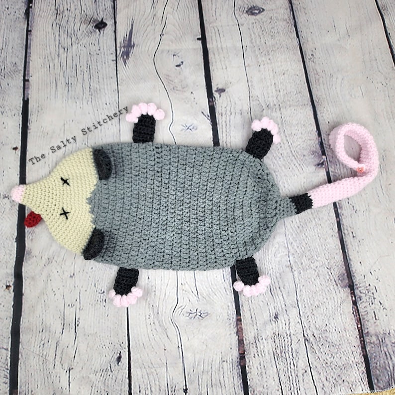 May include: A crocheted gray opossum with pink feet and a pink tail. The opossum has black eyes and a red tongue. The opossum is lying on its back with its legs in the air. The opossum is on a wooden background.