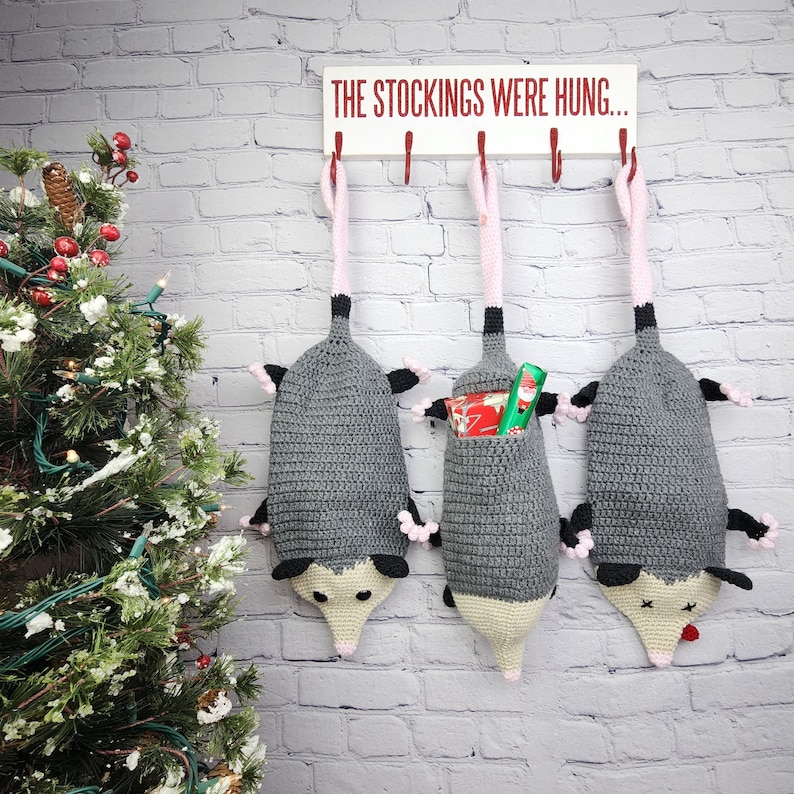 May include: Three gray crocheted opossum stockings hanging from hooks on a white brick wall. The stockings are filled with wrapped gifts and the sign above reads "The stockings were hung..."