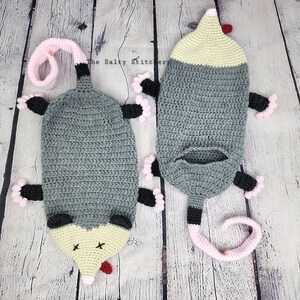 May include: Two crocheted opossum plushies, one lying down and one standing up. Both are gray with pink and black accents. The opossums have a yellow face with a red nose and black eyes.