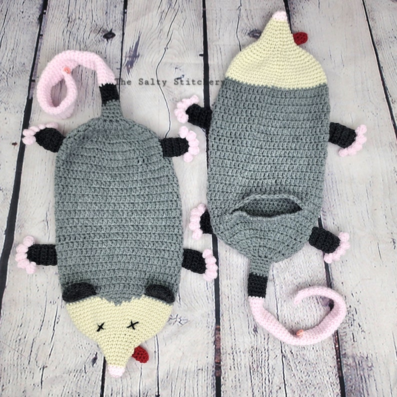 May include: Two crocheted opossum plushies, one lying down and one standing up. Both are gray with pink and black accents. The opossums have a yellow face with a red nose and black eyes.