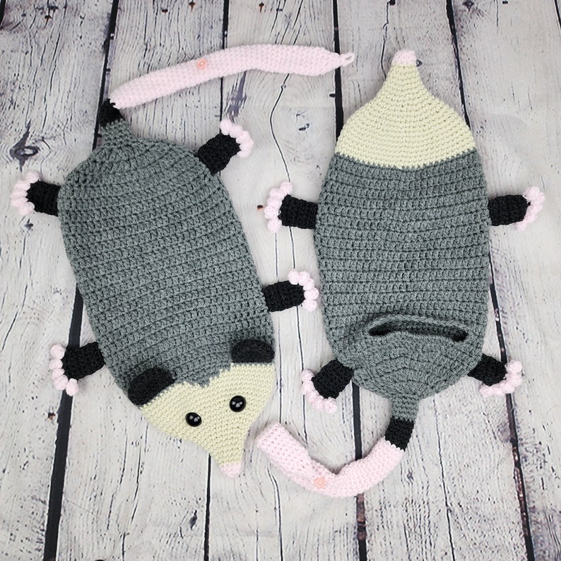 May include: Two crocheted opossum plushies with pink and black accents. The opossums are lying on a light wood surface.