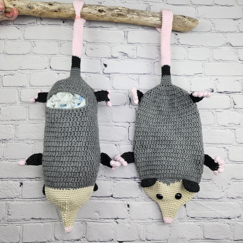 May include: Two gray crocheted opossum-shaped bags with pink ears and noses hanging from a wooden branch. The bags are designed to hold plastic bags.