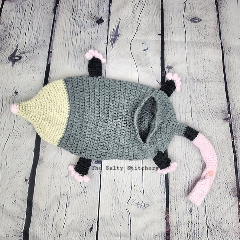 May include: A crocheted opossum costume in gray, pink, and black. The costume has a long tail and a pointed snout.