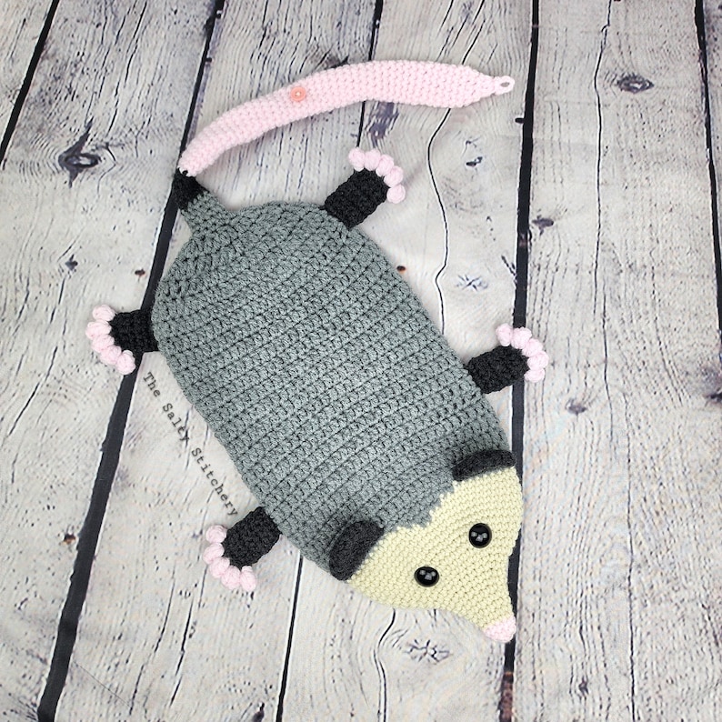 May include: A crocheted gray and yellow opossum plush toy with pink accents. The opossum has black eyes and a long tail. The text "The Salty Stitchery" is visible on the toy.