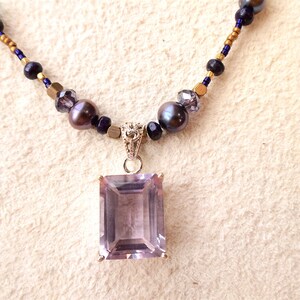 K&ouml;nnte beinhalten: Eine zarte Halskette mit einem gro&szlig;en, rechteckigen Amethyst-Anh&auml;nger. Die Halskette besteht aus kleinen, dunkelvioletten und goldenen Perlen.