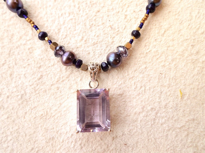 K&ouml;nnte beinhalten: Eine zarte Halskette mit einem gro&szlig;en, rechteckigen Amethyst-Anh&auml;nger. Die Halskette besteht aus kleinen, dunkelvioletten und goldenen Perlen.