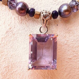 K&ouml;nnte beinhalten: Eine silberne Halskette mit einem Anh&auml;nger, der einen gro&szlig;en, rechteckigen, hellvioletten Amethyst-Edelstein tr&auml;gt. Die Halskette ist aus dunkelvioletten und goldenen Perlen gefertigt.