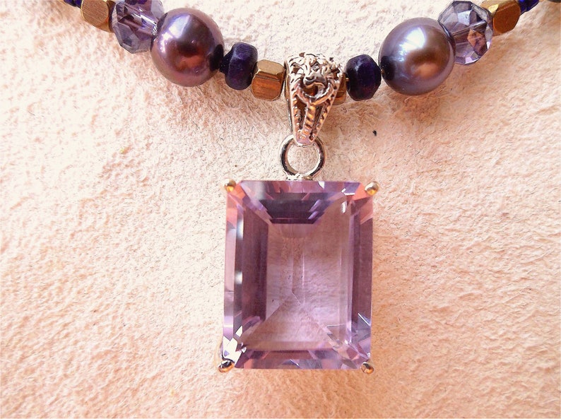 K&ouml;nnte beinhalten: Eine silberne Halskette mit einem Anh&auml;nger, der einen gro&szlig;en, rechteckigen, hellvioletten Amethyst-Edelstein tr&auml;gt. Die Halskette ist aus dunkelvioletten und goldenen Perlen gefertigt.