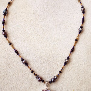 K&ouml;nnte beinhalten: Eine Halskette mit einem gro&szlig;en Amethyst-Anh&auml;nger. Die Halskette ist aus goldenen und schwarzen Perlen sowie kleinen Perlen gefertigt.