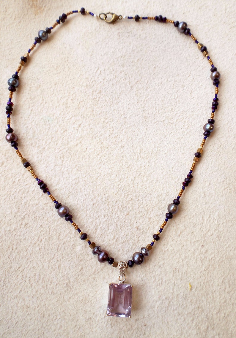 K&ouml;nnte beinhalten: Eine Halskette mit einem gro&szlig;en Amethyst-Anh&auml;nger. Die Halskette ist aus goldenen und schwarzen Perlen sowie kleinen Perlen gefertigt.