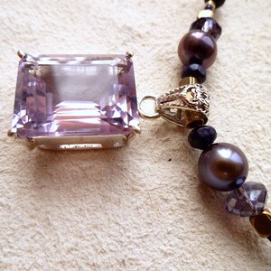 K&ouml;nnte beinhalten: Eine silberne Halskette mit einem gro&szlig;en, rechteckigen, hellvioletten Amethyst-Anh&auml;nger. Die Halskette besteht aus dunkelvioletten und goldenen Perlen.