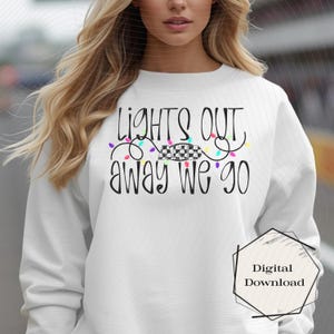 May include: White sweatshirt with the text "LIGHTS OUT AWAY WE GO" in black script. The design includes colorful string lights and a checkered flag. A digital download graphic is in the lower right corner.