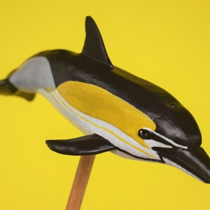 May include: A black and white dolphin figurine with yellow stripes, perched on a wooden stick against a yellow background.