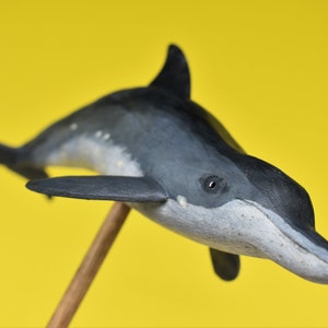 May include: A hand-painted figurine of a black and gray dolphin with a white underbelly. The dolphin is posed in a swimming position with its head tilted up. The figurine is mounted on a wooden stick.