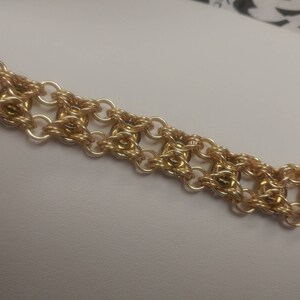 May include: A gold-colored bracelet with a chainmail design. The bracelet features a series of interconnected rings and small gold beads. The clasp is a simple bar and loop closure. The jewelry is displayed on a white surface.