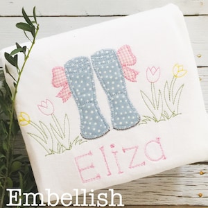 May include: A white embroidered t-shirt with a design of blue rain boots with pink bows, green grass, and yellow and pink tulips. The name "Eliza" is embroidered in pink below the design.