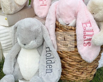 May include: A collection of plush bunny toys with personalized names embroidered on their ears. The bunnies are in various colors, including pink, gray, and white. The bunnies are sitting in a wicker basket.