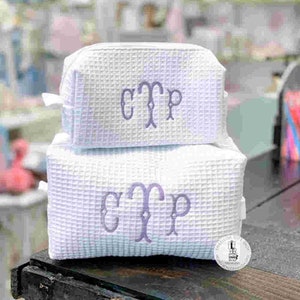 May include: Two white waffle-textured cosmetic bags stacked on top of each other. Each bag has a lavender monogram of the letters "CTP". The bags are rectangular with a zipper closure and a small loop on the side.