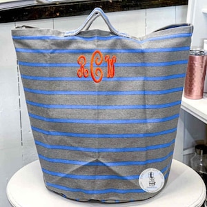 May include: A gray and blue striped canvas tote bag with a monogrammed "RCW" in orange thread. The bag has two handles and is sitting on a white surface.
