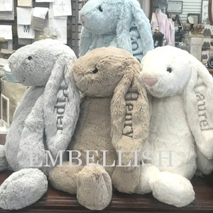 May include: Three plush bunny rabbits, one light blue, one light brown, and one white, are sitting on a wooden surface. Each bunny has a name embroidered on its ear: Piper, Henry, and Amelia.