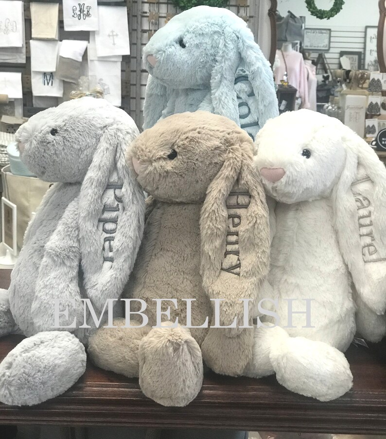 May include: Three plush bunny rabbits, one light blue, one light brown, and one white, are sitting on a wooden surface. Each bunny has a name embroidered on its ear: Piper, Henry, and Amelia.