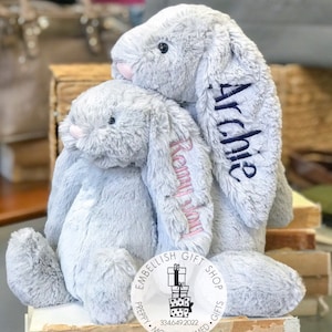 May include: Two gray plush bunny rabbits with embroidered names, "Remy" and "Archie", sitting together. The bunnies are from a shop called "Embellish Gift Shop - Preppy Monogrammed Gifts".