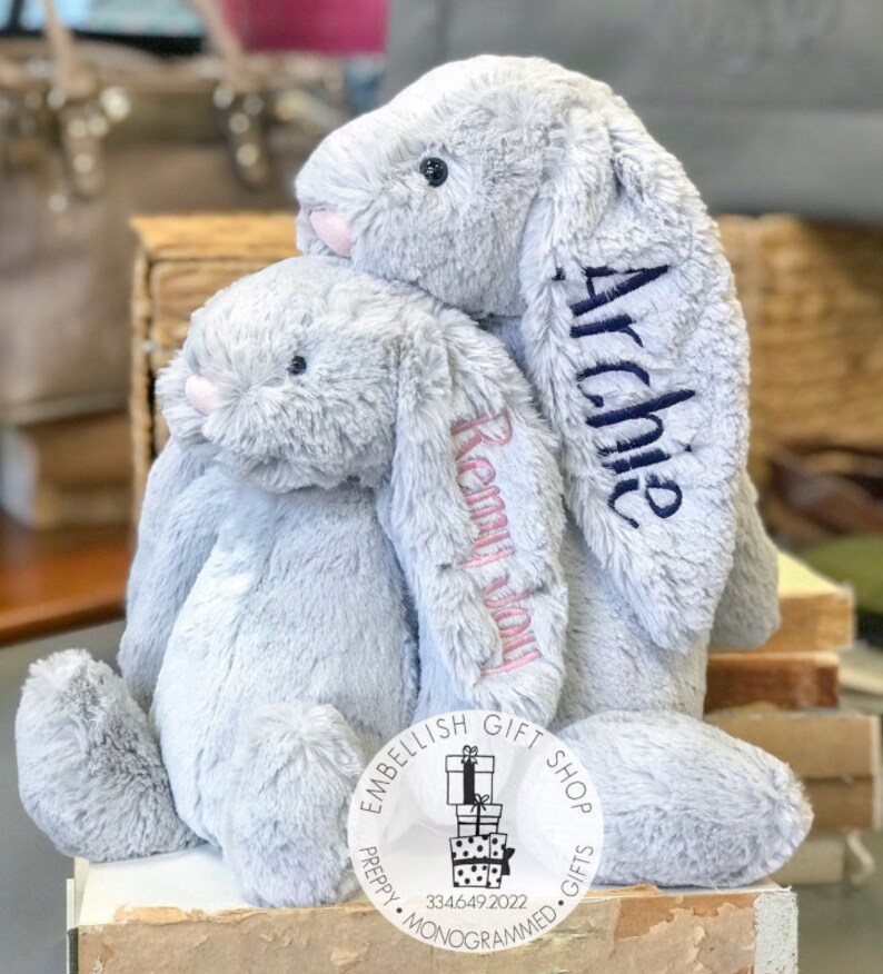 May include: Two gray plush bunny rabbits with embroidered names, "Remy" and "Archie", sitting together. The bunnies are from a shop called "Embellish Gift Shop - Preppy Monogrammed Gifts".