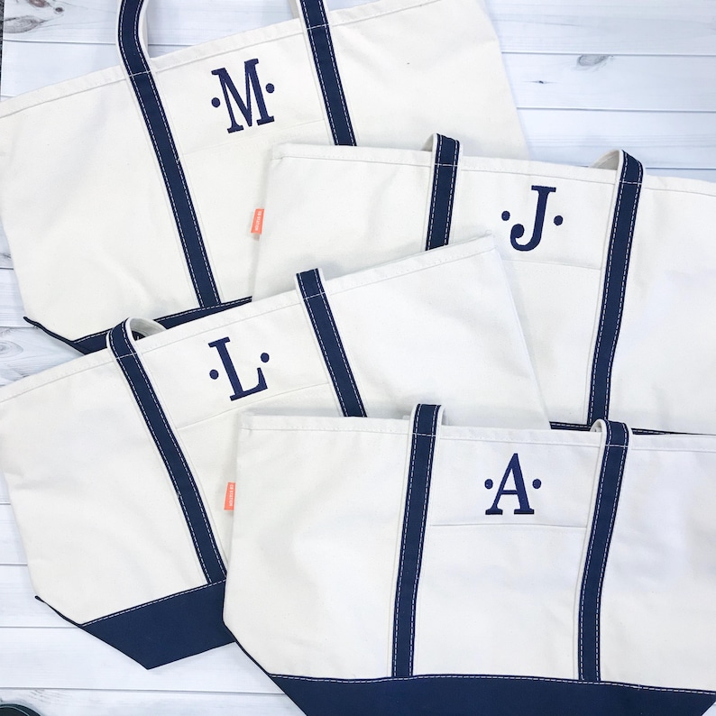 May include: Four white canvas tote bags with navy blue handles and trim. Each bag has a single letter monogrammed in navy blue on the front. The letters are M, J, L, and A.
