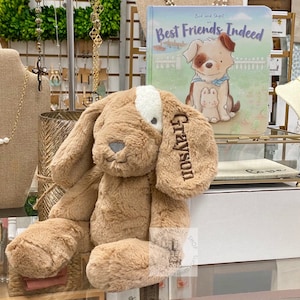 May include: A brown plush dog toy with a white patch on its chest. The dog is sitting on a table and has the name "Grayson" embroidered on its ear. The toy is next to a book titled "Best Friends Indeed" with a picture of two dogs on the cover.
